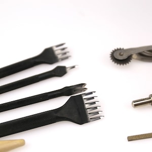 May include: A collection of leatherworking tools, including several black stitching chisels with textured handles, a rotary leather edge creaser, a metal edge beveler, and a small metal tool with a right-angle end. The tools are arranged on a white surface.