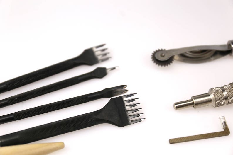 May include: A collection of leatherworking tools, including several black stitching chisels with textured handles, a rotary leather edge creaser, a metal edge beveler, and a small metal tool with a right-angle end. The tools are arranged on a white surface.