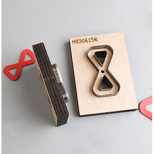 Pu&ograve; includere: Un modello di fustellatura in legno con il numero "M030615R" stampato su di esso. Il modello ha la forma di un simbolo di infinito con un simbolo di infinito pi&ugrave; piccolo all'interno. Il modello &egrave; su una superficie grigia.