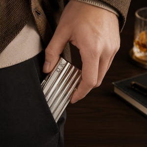 May include: A silver metal cigarette case with a ribbed design is being placed into a black pocket. The case has the initials "D.K." engraved on the top. The person is wearing a brown blazer and a beige sweater. A glass of amber liquid and a notebook are in the background.