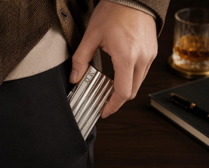 May include: A silver metal cigarette case with a ribbed design is being placed into a black pocket. The case has the initials "D.K." engraved on the top. The person is wearing a brown blazer and a beige sweater. A glass of amber liquid and a notebook are in the background.