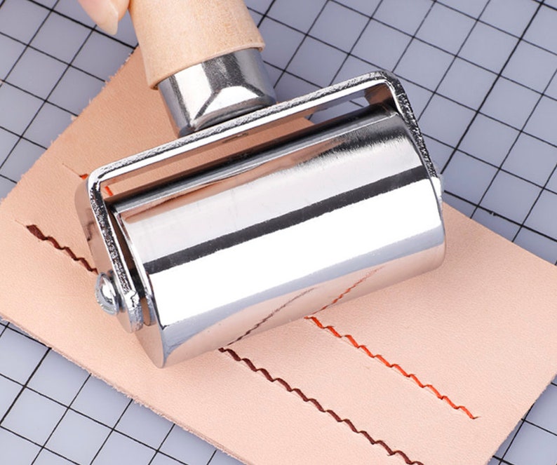 May include: A metal leathercraft roller with a wooden handle is pressed onto a piece of light brown leather. The roller has a silver finish and is used to create a decorative edge. The leather has a grid pattern in the background.