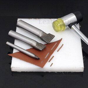 May include: A set of four leather crafting tools, including three different sized punches and a mallet with a yellow rubber head. The tools are on a white cutting mat with a piece of brown leather.