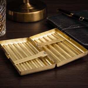 May include: Open, gold-colored cigarette case with a ribbed interior. The case is rectangular and has a hinged lid. A black leather notebook and pen are in the background. A glass of amber liquid is visible on the left.