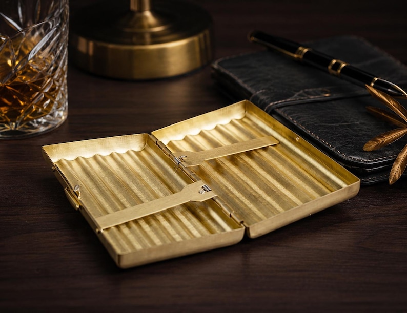 May include: Open, gold-colored cigarette case with a ribbed interior. The case is rectangular and has a hinged lid. A black leather notebook and pen are in the background. A glass of amber liquid is visible on the left.