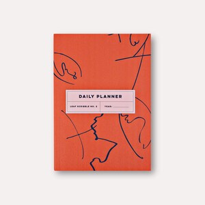 Puede incluir: Una agenda diaria rectangular cerrada con una cubierta naranja con arte lineal abstracto en azul marino. La agenda tiene una etiqueta rectangular que dice "DAILY PLANNER" y "LEAF SCRIBBLE NO. 2 YEAR:"