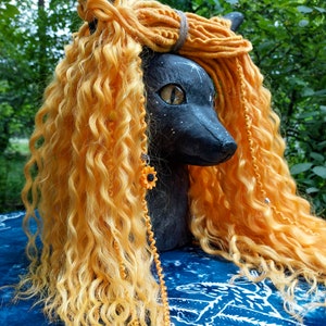 May include: A black cat sculpture with yellow curly hair and dreadlocks. The cat has yellow eyes and a sunflower earring. The sculpture is set against a blue and white patterned background, with green foliage in the background.