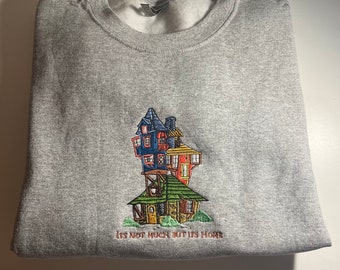 May include: A heather gray crewneck sweatshirt featuring a detailed embroidered graphic of a multi-story, whimsical house with blue, yellow, red, and green sections. Below the house, the text reads, "ITS NOT MUCH BUT ITS HOME" in brown thread.