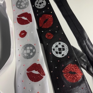 May include: Two satin neckties, one white and one black, each decorated with a repeating pattern of glittery silver disco balls and red glittery lips. The ties are embellished with small, scattered rhinestones in clear and light pink tones.