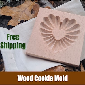 May include: A wooden cookie mold shaped like a heart with a sunburst design. The mold is light brown and has a smooth surface. Free Shipping is written on a white fabric bag behind the mold.