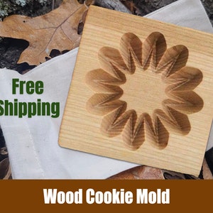 May include: A wooden cookie mold with a flower design. The mold is light brown and has a smooth surface. The mold is sitting on a white cloth with the words "Free Shipping" printed on it.
