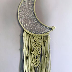 May include: A handmade, moon-shaped dream catcher in shades of green. The crescent frame is adorned with a delicate web and small, light green beads. The lower portion features intricate macrame knotting and hanging cords.