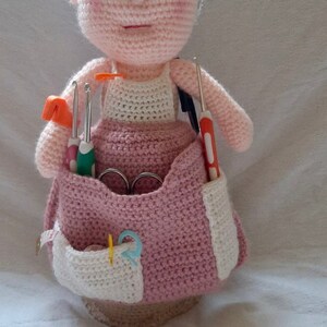 Puede incluir: Una muñeca de crochet con un vestido rosa y un delantal blanco, sosteniendo una variedad de agujas de crochet y tijeras en una bolsa de crochet rosa y blanca.