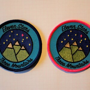 Könnte beinhalten: Zwei gestickte Patches mit blauem Hintergrund, grünen Bergen und gelben Sternen. Der Text auf den Patches lautet "Eleven Stars Three Mountains". Ein Patch hat einen schwarzen Rand und der andere einen rosa Rand.