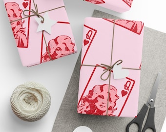 May include: Two rectangular gifts wrapped in light pink paper featuring a repeating red graphic of a woman's face in the style of a playing card queen. Each package is tied with brown twine and adorned with a white star or heart-shaped tag.