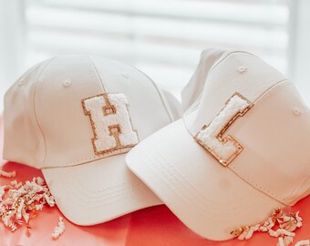 May include: Two white baseball caps with gold glitter letters. The first cap has a large "H" on the front and the second cap has a large "L" on the front.