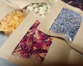 May include: Six small brown paper bags with clear plastic windows, each filled with a different type of dried flower. The flowers include lavender, rose petals, jasmine, orange blossoms, and hibiscus.