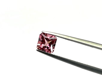May include: A square-cut, pink gemstone is held by silver tweezers against a white background. The gemstone has a faceted surface, reflecting light. The tweezers cast a shadow, adding depth to the image. The gem is a vibrant shade of pink.