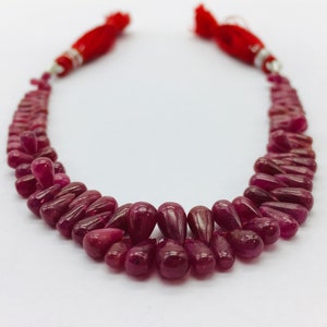 Peut inclure: Un collier composé de nombreuses perles en forme de goutte, d'un rouge profond. Les perles sont enfilées ensemble, formant une forme incurvée. Le collier est orné de pompons rouges à chaque extrémité.