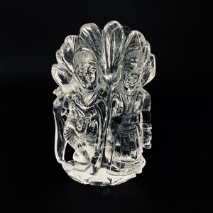 May include: A clear quartz sculpture of two figures standing together. The figures are carved in a traditional style and are surrounded by a decorative floral design.
