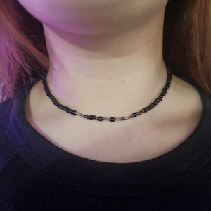 May include: A black beaded choker necklace. The necklace is made of small, dark beads, with a few larger beads and metallic accents. The necklace is worn around the neck, against a black top.