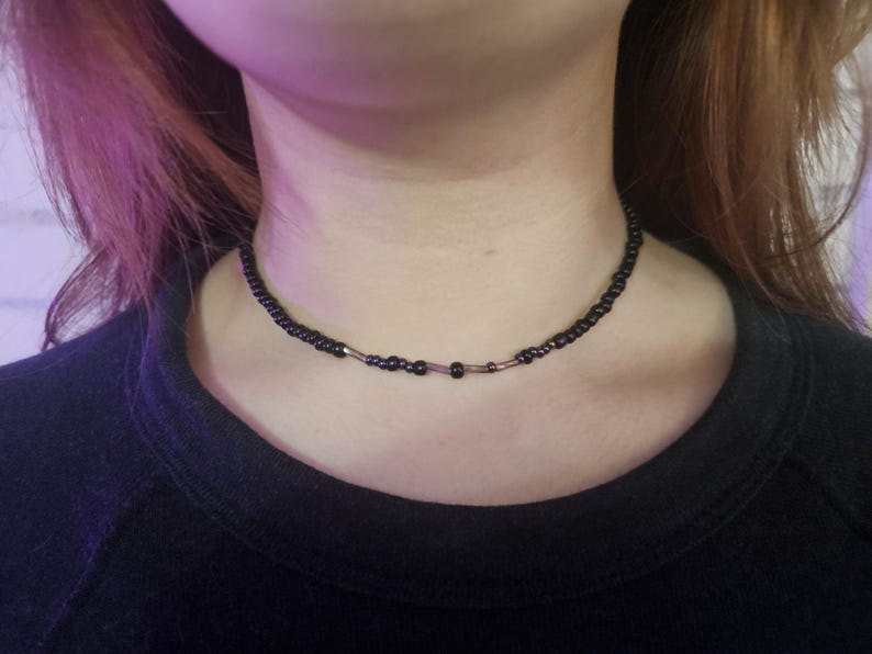 May include: A black beaded choker necklace. The necklace is made of small, dark beads, with a few larger beads and metallic accents. The necklace is worn around the neck, against a black top.
