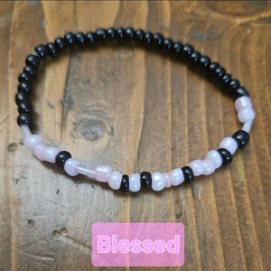 Peut inclure: Un bracelet de perles noires et roses p&acirc;les. Le bracelet pr&eacute;sente un motif en spirale. Le mot "Blessed" est affich&eacute; sur un fond rose.