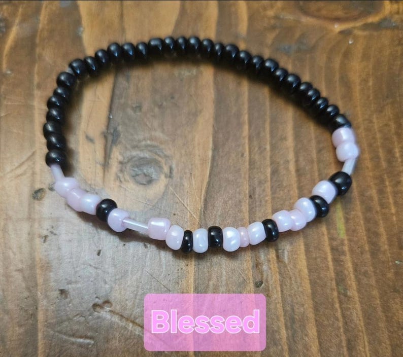Peut inclure: Un bracelet de perles noires et roses p&acirc;les. Le bracelet pr&eacute;sente un motif en spirale. Le mot "Blessed" est affich&eacute; sur un fond rose.