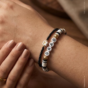May include: Two beaded bracelets on a wrist. One is black with gold beads, the other is white and black with gold accents. The word "GRAD" is spelled out on the white and black bracelet. The text "THE COLLECTION / GRADUATION '24" is visible.