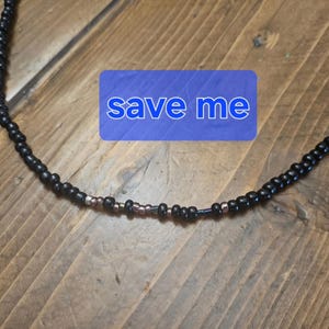 May include: A black beaded necklace with a blue rectangle bearing the words "save me". The necklace features a combination of black and copper-coloured beads. It is presented on a wooden surface.