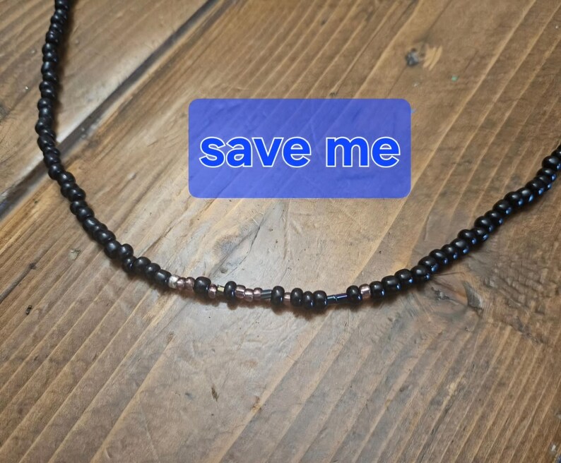 May include: A black beaded necklace with a blue rectangle bearing the words "save me". The necklace features a combination of black and copper-coloured beads. It is presented on a wooden surface.