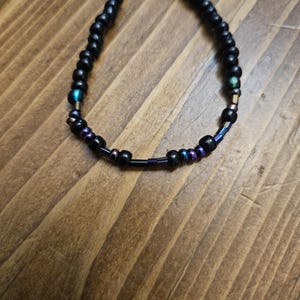 Peut inclure: Bracelet de perles noires avec des accents iris&eacute;s. Le bracelet est compos&eacute; de petites perles noires rondes, avec des perles iris&eacute;es bleues, violettes et vertes. Le bracelet est pr&eacute;sent&eacute; sur une surface en bois.