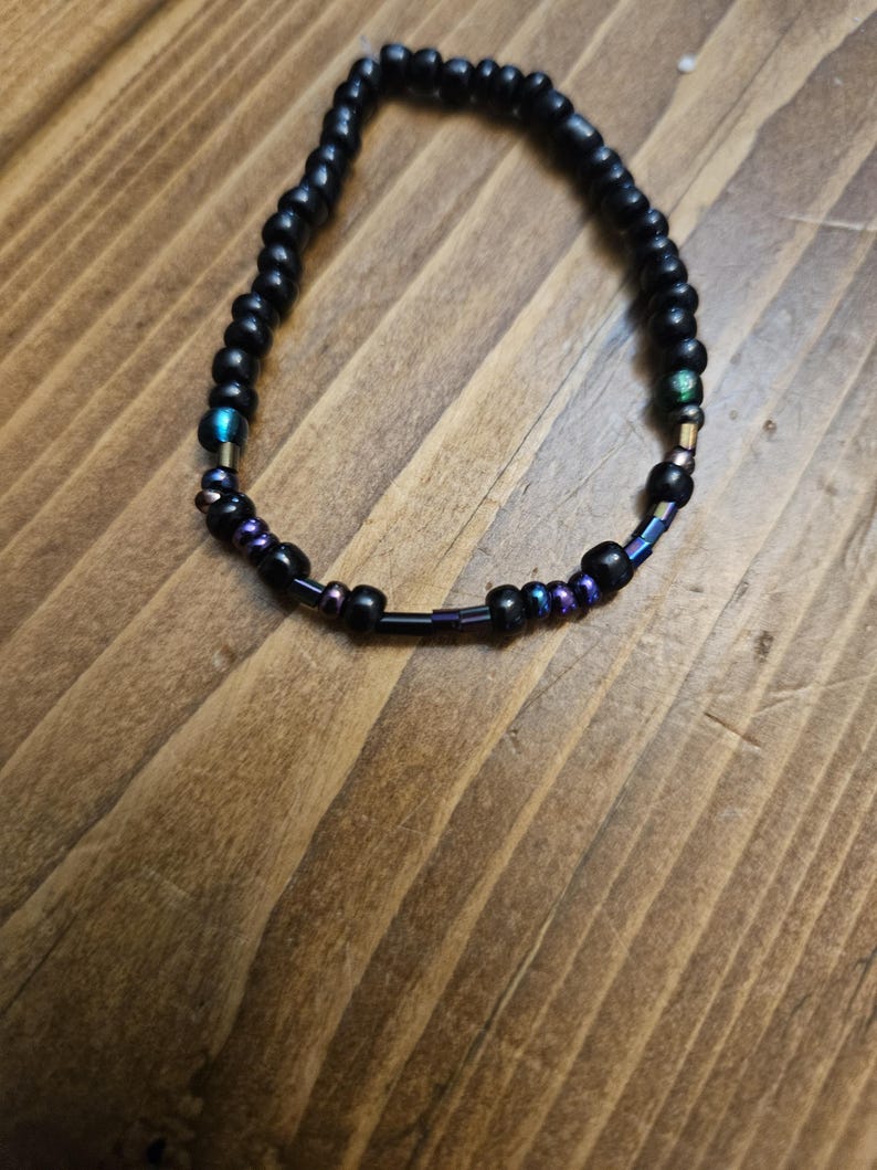 Peut inclure: Bracelet de perles noires avec des accents iris&eacute;s. Le bracelet est compos&eacute; de petites perles noires rondes, avec des perles iris&eacute;es bleues, violettes et vertes. Le bracelet est pr&eacute;sent&eacute; sur une surface en bois.