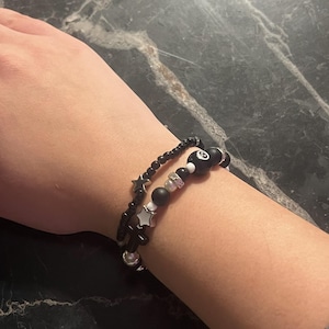 May include: Two beaded bracelets in black and white. One bracelet features small black beads and silver star charms. The other bracelet has black beads, white beads, and an 8-ball charm. The bracelets are worn on a wrist.