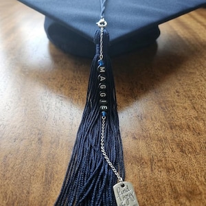 May include: A navy blue graduation cap with a matching tassel. The tassel features the name "MAGGIE" in beads and a silver charm inscribed with "Find Joy in the Journey."