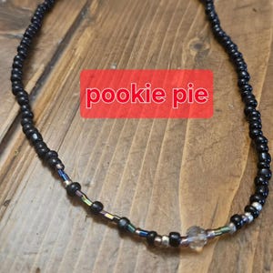 May include: A black beaded necklace with iridescent beads and a clear, faceted bead. The necklace has the text "pookie pie" on a red background. The beads are small and evenly spaced, creating a simple design.