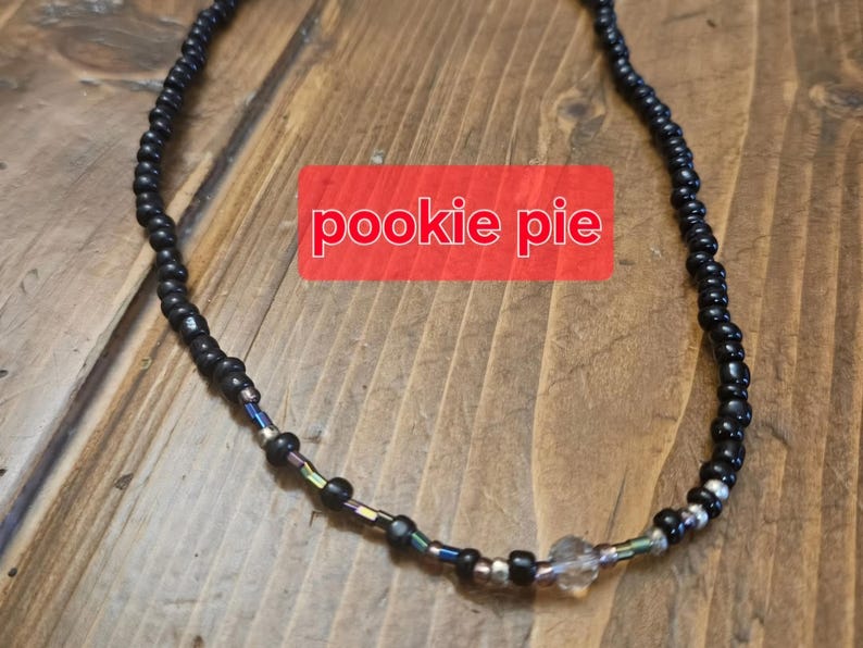 May include: A black beaded necklace with iridescent beads and a clear, faceted bead. The necklace has the text "pookie pie" on a red background. The beads are small and evenly spaced, creating a simple design.