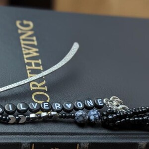 May include: A silver metal bookmark with a beaded charm that spells out "SILVERONE". The charm includes black beads, silver accents, and a decorative tassel. The bookmark rests on a dark book with gold lettering.