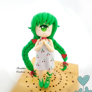 May include: A handmade figurine of a girl with green hair and eyes, wearing a white dress with red cherry designs. The figurine has green shoes and long green braids with red heart accents. The figurine is on a wooden surface. The text "Floratales Creations" is visible.