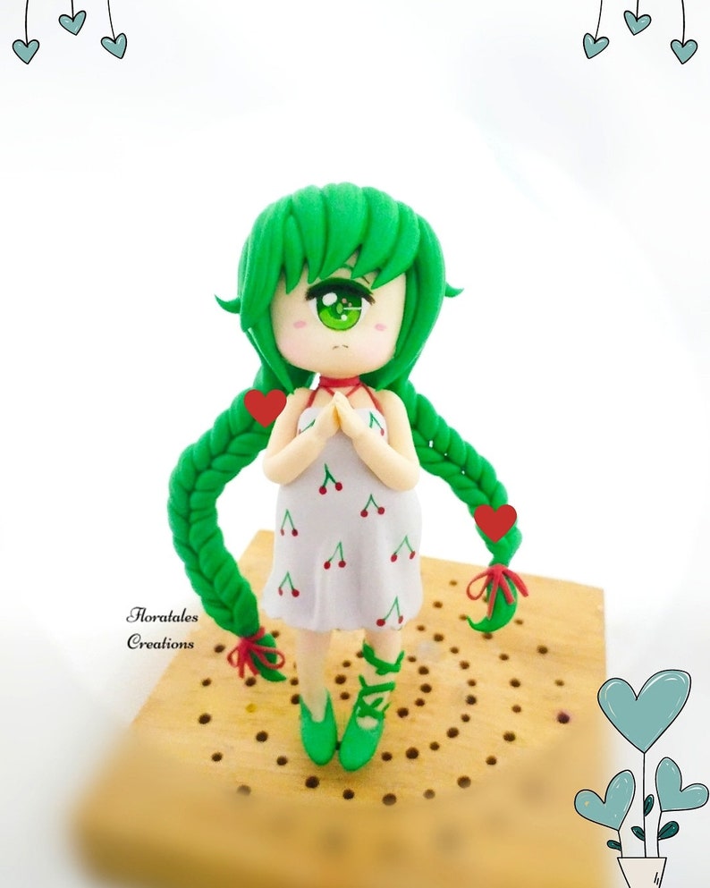 May include: A handmade figurine of a girl with green hair and eyes, wearing a white dress with red cherry designs. The figurine has green shoes and long green braids with red heart accents. The figurine is on a wooden surface. The text "Floratales Creations" is visible.
