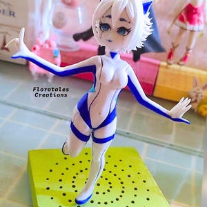 K&ouml;nnte beinhalten: Eine wei&szlig;e und blaue Figur einer weiblichen Figur mit Katzenohren. Die Figur ist mit einem erhobenen Bein und einem ausgestreckten Arm posiert. Die Figur steht auf einem gr&uuml;nen Sockel mit L&ouml;chern.