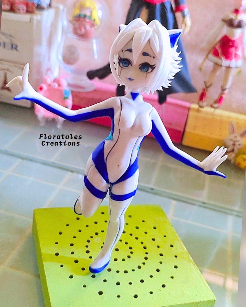 K&ouml;nnte beinhalten: Eine wei&szlig;e und blaue Figur einer weiblichen Figur mit Katzenohren. Die Figur ist mit einem erhobenen Bein und einem ausgestreckten Arm posiert. Die Figur steht auf einem gr&uuml;nen Sockel mit L&ouml;chern.