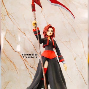 May include: A figurine of a woman in a black and red outfit holding a red scythe. The figurine has red hair and is standing in front of a marble-like background. The text "Floratales Creations" is visible.