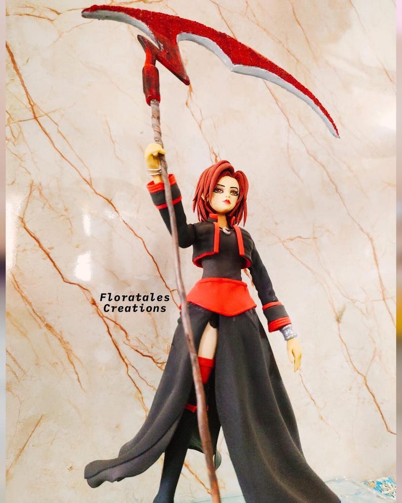 May include: A figurine of a woman in a black and red outfit holding a red scythe. The figurine has red hair and is standing in front of a marble-like background. The text "Floratales Creations" is visible.