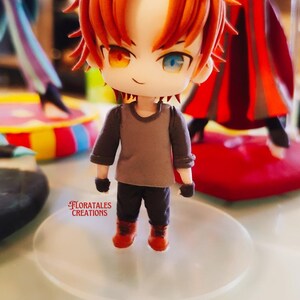 May include: A small figurine of a character with orange hair and mismatched eyes, one orange and one blue. The figure wears a gray shirt, black pants, and orange boots. The base is clear, and the text "FLORATALES CREATIONS" is visible.