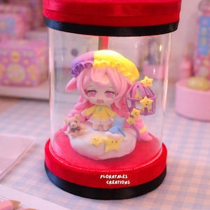 May include: A miniature figurine of a character with pink hair and a yellow dress, inside a clear cylindrical container with a red base and lid. The figurine is surrounded by stars and a small teddy bear. The text "FLORATALES CREATIONS" is visible.
