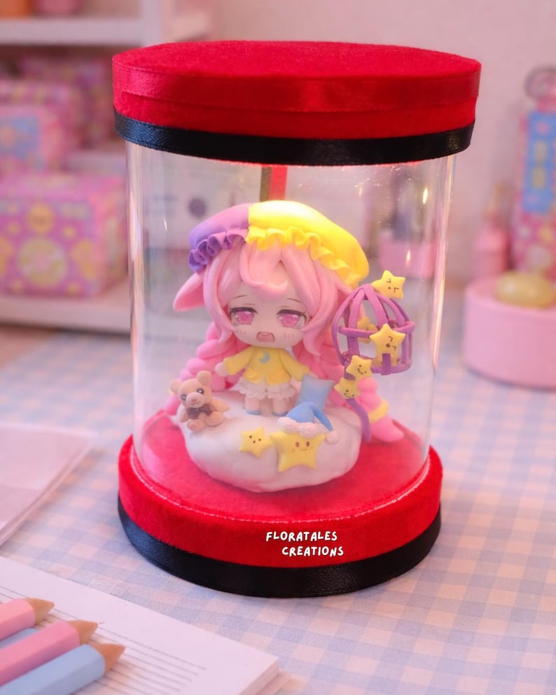 May include: A miniature figurine of a character with pink hair and a yellow dress, inside a clear cylindrical container with a red base and lid. The figurine is surrounded by stars and a small teddy bear. The text "FLORATALES CREATIONS" is visible.