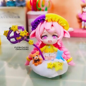 May include: A handcrafted clay figurine of a character with pink hair, a yellow and purple hat, and a yellow dress. The figurine is holding a purple wand with yellow star accents. A small teddy bear and stars are on a cloud base. The text "FLORATALES CREATIONS" is visible.