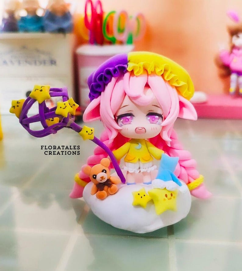 May include: A handcrafted clay figurine of a character with pink hair, a yellow and purple hat, and a yellow dress. The figurine is holding a purple wand with yellow star accents. A small teddy bear and stars are on a cloud base. The text "FLORATALES CREATIONS" is visible.