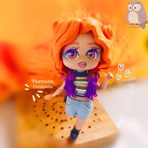 May include: A handmade doll with orange hair, blue eyes, and a striped shirt. The doll is wearing blue shorts and black shoes. The text "Floratales Creations" is visible. A cartoon owl is in the background.